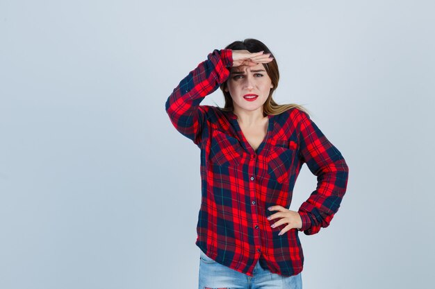 Retrato de mujer joven mirando al frente con las manos sobre la cabeza en camisa a cuadros y mirando confundido vista frontal