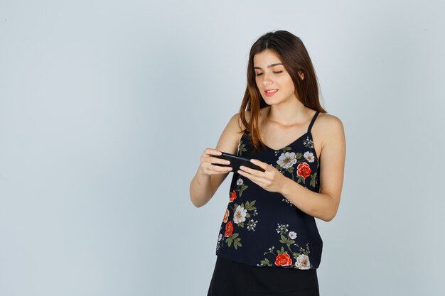Retrato de mujer joven mirando smartphone en blusa, falda y mirando alegre vista frontal
