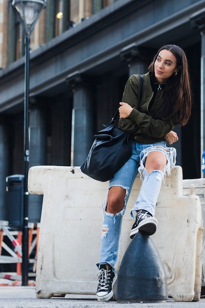 Retrato de una mujer joven de moda con el bolso que se sienta en la calle
