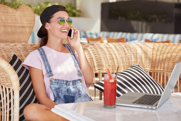 Retrato de mujer joven de moda feliz con gafas de sol y gorra negra, se sienta contra el interior del café, disfruta de la conversación móvil, trabaja en una computadora portátil, comparte noticias positivas con su mejor amigo