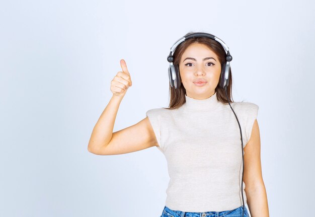 Retrato de mujer joven modelo escuchando música en auriculares y mostrando un pulgar hacia arriba.