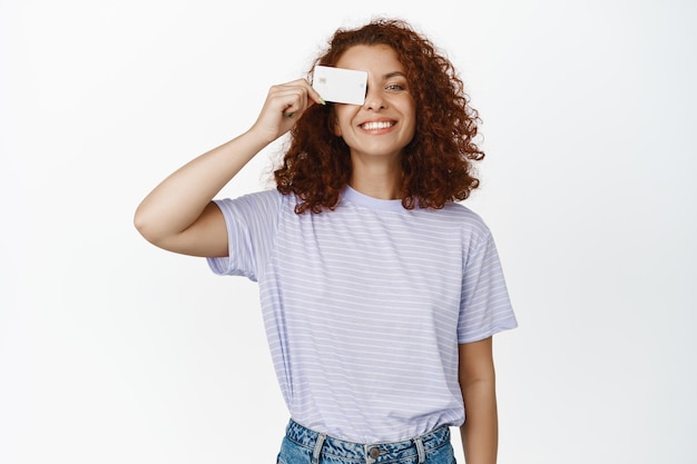 Retrato de mujer joven moderna que muestra la tarjeta de crédito cerca de la cara, sonriendo feliz, pagando sin contacto, le gusta ir de compras, fondo blanco