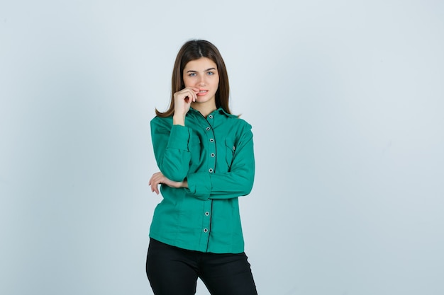 Retrato de mujer joven mordiéndose las uñas en camisa verde, pantalones y mirando pensativo vista frontal