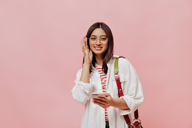 Retrato de mujer joven morena bronceada en anteojos y camisa blanca sostiene teléfono celular y sonríe sobre fondo rosa aislado