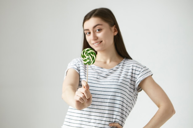 Retrato de mujer joven morena hermosa alegre en camiseta casual rayada con paleta, sonriendo con expresión facial alegre positiva. Enfoque selectivo en caramelos duros