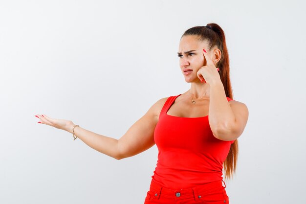 Retrato de mujer joven mostrando algo en camiseta roja, pantalones y mirando pensativo vista frontal