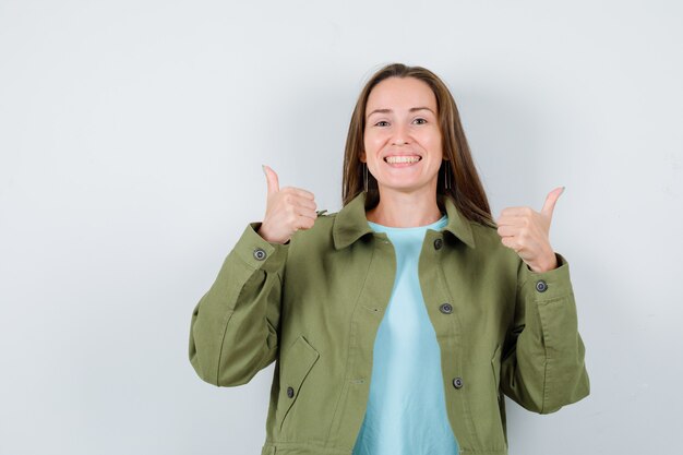 Retrato de mujer joven mostrando doble pulgar hacia arriba en camiseta, chaqueta y mirando alegre vista frontal