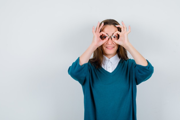 Retrato de mujer joven mostrando gesto de gafas en suéter sobre camisa y mirando sorprendido vista frontal