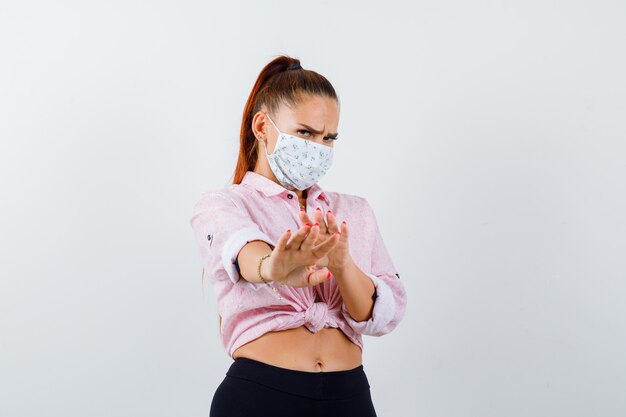 Retrato de mujer joven mostrando gesto de parada en camisa, pantalón, máscara médica y mirando asustado vista frontal