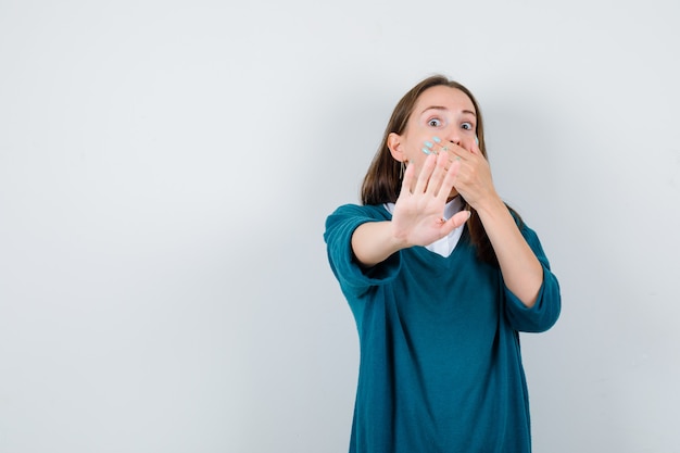 Retrato de mujer joven mostrando gesto de parada, manteniendo la mano en la boca en suéter sobre camisa blanca y mirando aterrorizado vista frontal
