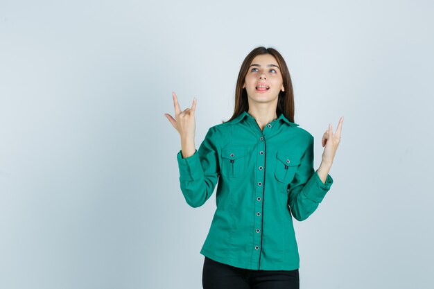 Retrato de mujer joven mostrando gesto de rock, sacando la lengua en camisa verde y mirando feliz vista frontal