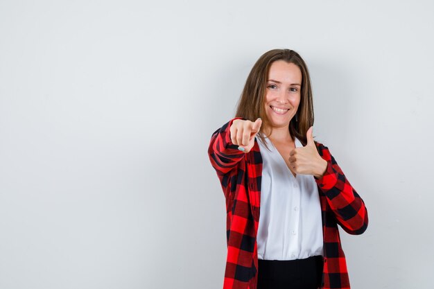 Retrato de mujer joven mostrando el pulgar hacia arriba, apuntando hacia adelante en ropa casual y mirando alegre vista frontal