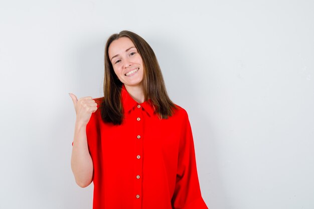 Retrato de mujer joven mostrando el pulgar hacia arriba en blusa roja y mirando alegre vista frontal