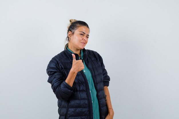 Retrato de mujer joven mostrando el pulgar hacia arriba en camisa, chaqueta acolchada y mirando afortunado vista frontal