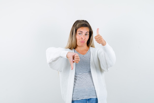 Retrato de mujer joven mostrando los pulgares hacia arriba y hacia abajo en camiseta, chaqueta y mirando vacilante vista frontal