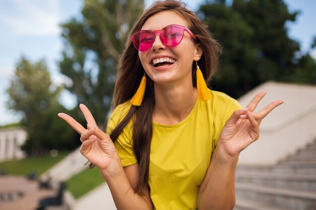 Retrato de mujer joven muy sonriente divirtiéndose en el parque de la ciudad, positiva, emocional, con top amarillo, aretes, gafas de sol rosas, tendencia de moda de estilo veraniego, accesorios elegantes, mostrando el signo de la paz