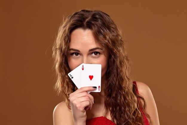 Retrato de una mujer joven o de cabello castaño sonriendo, sosteniendo un par de ases con un vestido de cóctel rojo sobre fondo marrón. Concepto de casino, industria del juego