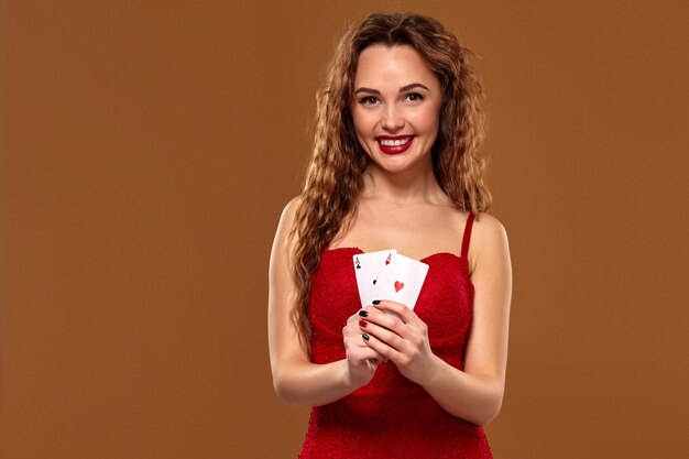 Retrato de una mujer joven o de cabello castaño sonriendo, sosteniendo un par de ases con un vestido de cóctel rojo sobre fondo marrón. Concepto de casino, industria del juego