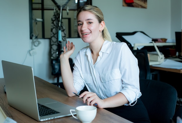Retrato de mujer joven oficinista sentada en el escritorio de oficina con ordenador portátil mirando a la cámara sonriendo alegremente trabajando en la oficina