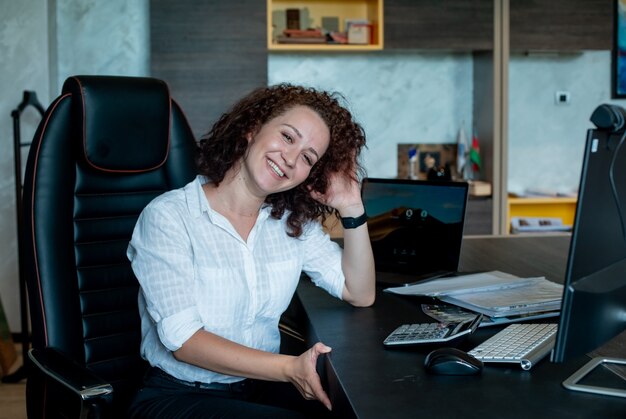 Retrato de mujer joven oficinista sentada en el escritorio de oficina con ordenador portátil mirando a la cámara sonriendo alegremente trabajando en la oficina