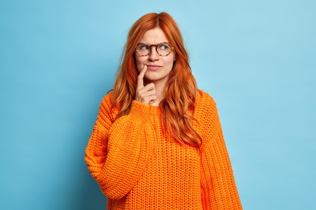 Retrato de mujer joven pelirroja atractiva mantiene el dedo índice cerca de la esquina de los labios, piensa demasiado en algo y toma la decisión de usar suéter de punto.