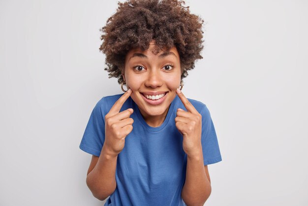 Retrato de mujer joven de pelo rizado alegre señala con el dedo índice en las mejillas sonríe ampliamente se siente muy feliz vestida con camiseta azul casual aislada sobre fondo blanco. Mira mi cara feliz