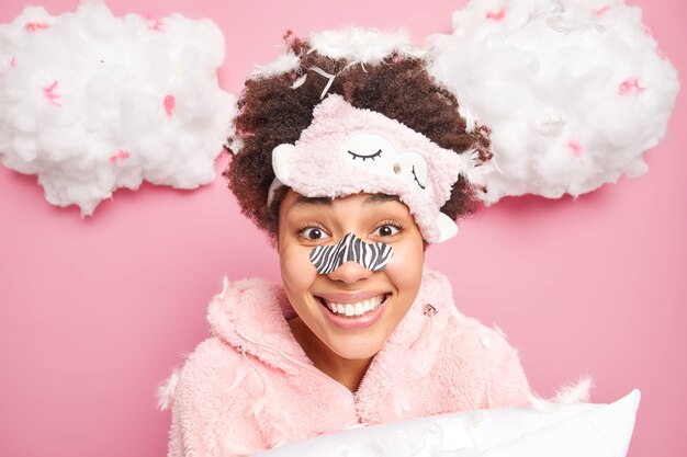 Retrato de mujer joven de pelo rizado feliz aplica ruta de eliminación de puntos negros en la nariz sonríe ampliamente se somete a procedimientos de belleza para el cuidado de la piel vestida con poses de pijama con almohada y plumas voladoras