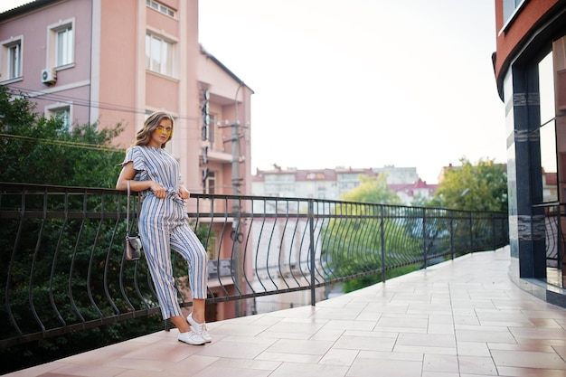 Retrato de una mujer joven perfecta con gafas de sol amarillas y a rayas posa con su bolso en un balcón de un edificio moderno en una ciudad