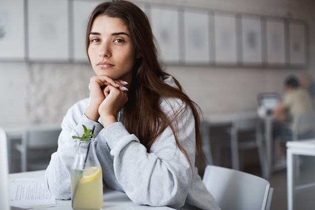 Retrato de mujer joven perfecta en sudadera gris bebiendo limonada en la gran biblioteca de espacios abiertos.