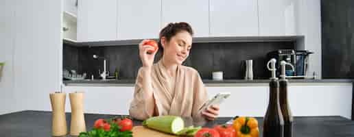 Foto gratuita retrato de una mujer joven pidiendo comestibles en una aplicación para teléfonos inteligentes sosteniendo un tomate sentado cerca de cortar