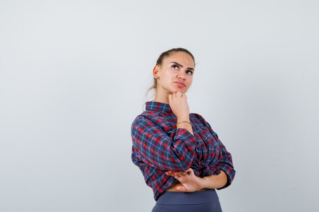 Retrato de mujer joven de pie en pose de pensamiento en camisa a cuadros, pantalones y mirando pensativo vista frontal