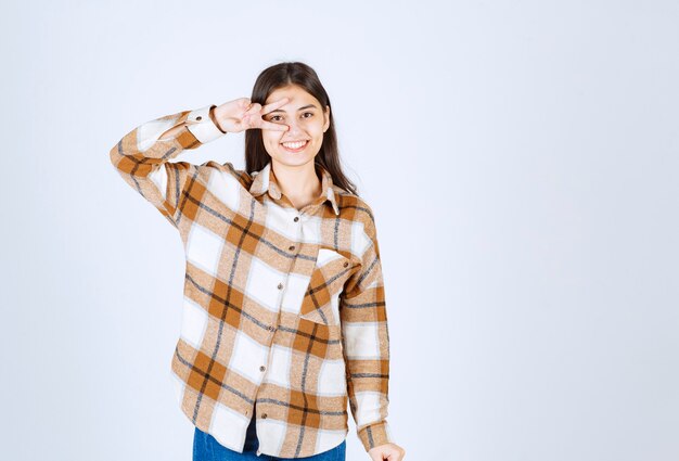 Retrato de mujer joven de pie y sonriendo a la cámara sobre una pared blanca.