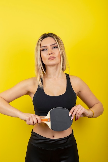 Retrato de mujer joven de pie y sosteniendo la raqueta de tenis de mesa en la pared amarilla.