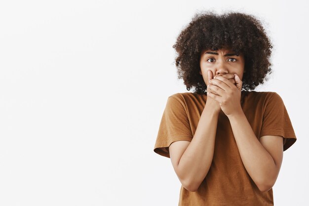 Retrato de mujer joven de piel oscura molesta preocupada con peinado afro cubriendo la boca con ambas palmas frunciendo el ceño, lo siento por decir palabras groseras