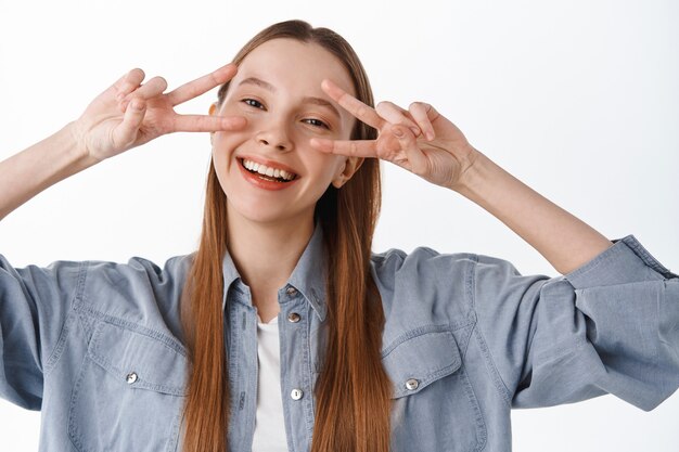 Retrato de mujer joven positiva de cerca, mostrando un gesto de paz con signo de v y sonriendo feliz con dientes blancos, de pie optimista contra la pared del estudio