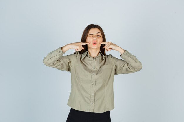 Foto gratuita retrato de mujer joven presionando los dedos en las mejillas mientras fruncía el ceño en camisa