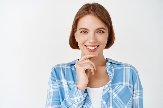 Retrato de mujer joven profesional con sonrisa segura, tocando la barbilla y mirando, de pie sobre la pared blanca