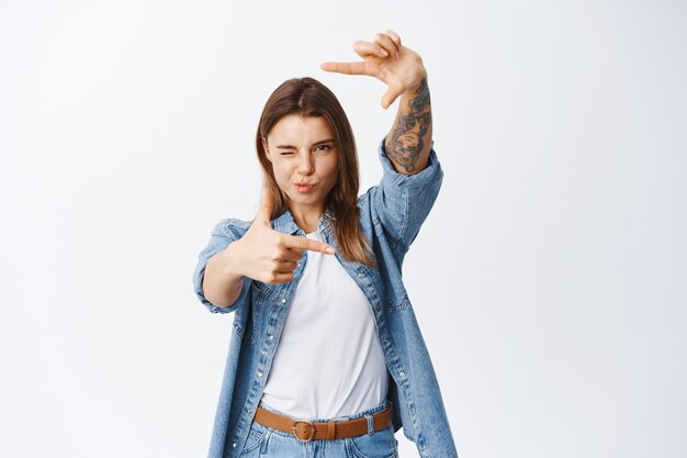 Retrato de mujer joven que busca el ángulo perfecto, haciendo marcos de dedos de la cámara y mirando a través de ella, de pie sobre una pared blanca