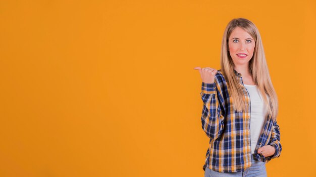 Retrato de una mujer joven que mira y señala al lado con el pulgar hacia arriba sobre un fondo naranja