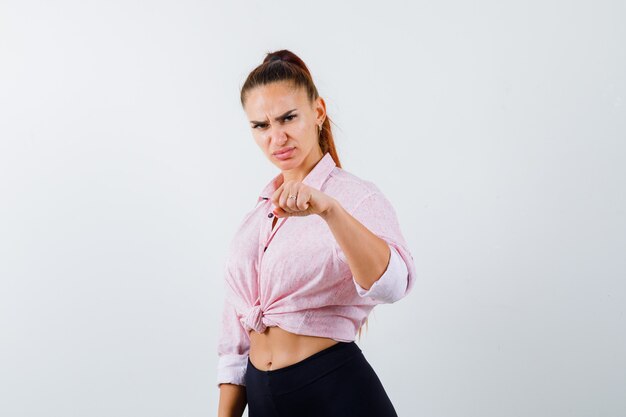 Retrato de mujer joven que muestra el puño cerrado en camisa casual, pantalones y mirando seriamente vista frontal