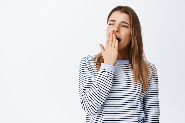 Retrato de mujer joven que se siente cansada, bostezando y cubriendo la boca abierta con la mano, de pie, somnolienta o aburrida, de pie sobre blanco