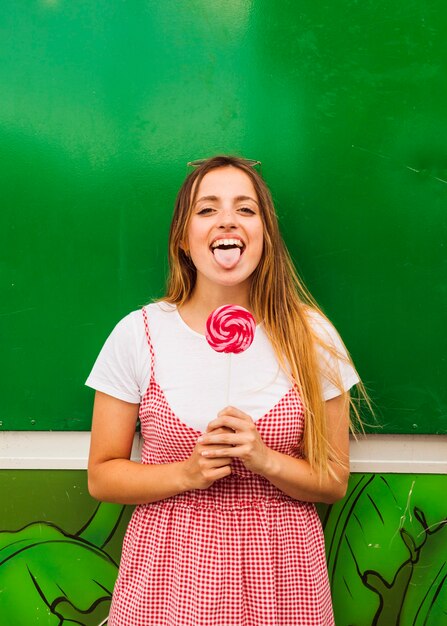 Retrato de una mujer joven que sostiene la piruleta roja en la mano que muestra su lengua