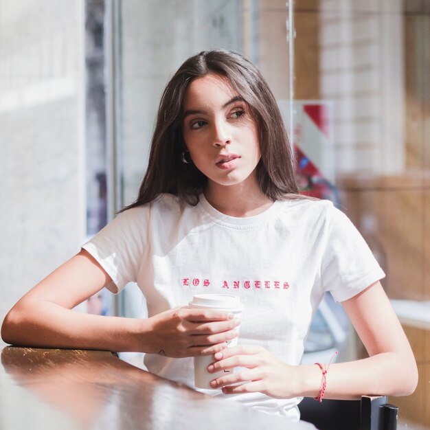 Retrato de una mujer joven que sostiene la taza de café disponible en la mano que se sienta en el café