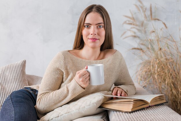 Retrato de una mujer joven que sostiene la taza de café que se sienta en el sofá con el libro