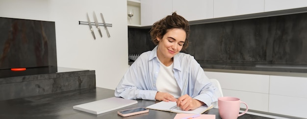 Foto gratuita retrato de una mujer joven que trabaja desde casa escribiendo información en un cuaderno tomando notas sentada