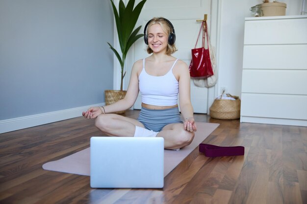Foto gratuita retrato de una mujer joven y relajada usando una computadora portátil y auriculares inalámbricos mientras practica la meditación