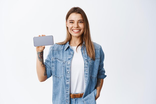 Retrato de mujer joven en ropa casual sosteniendo el teléfono inteligente horizontal, mostrando la pantalla vacía del teléfono móvil para el anuncio de la aplicación, blanco