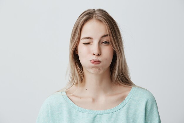 Retrato de mujer joven rubia con el ceño fruncido haciendo pucheros en las mejillas, parpadeando