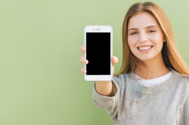 Retrato de una mujer joven rubia feliz que muestra el teléfono móvil contra el contexto verde