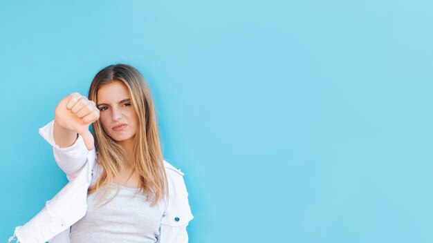 Retrato de una mujer joven rubia nerviosa que muestra el pulgar hacia abajo contra el fondo azul